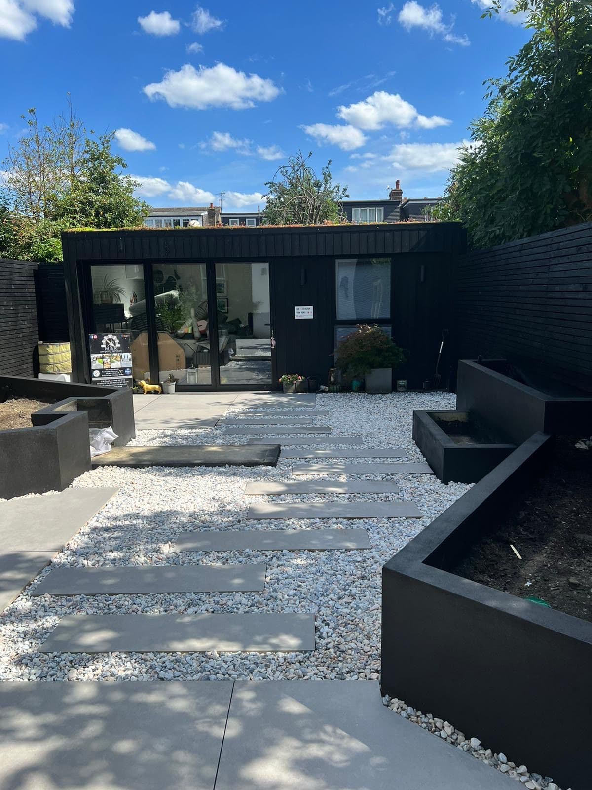Compact landscaped garden by M&R Property & Paving Services with stepping slabs, black raised planters and a garden room